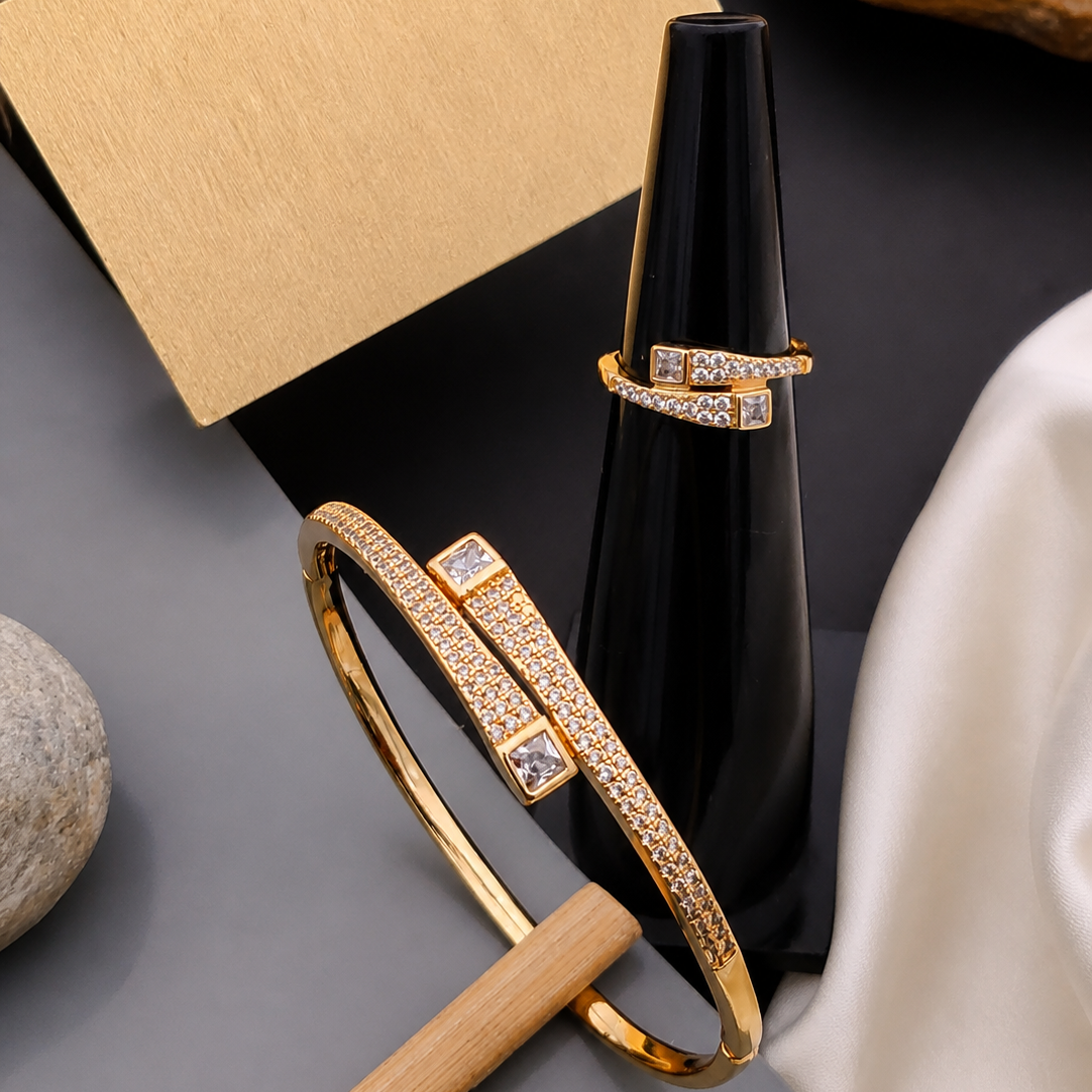 Gold bracelet and ring on a dark surface with a black vase and beige box.